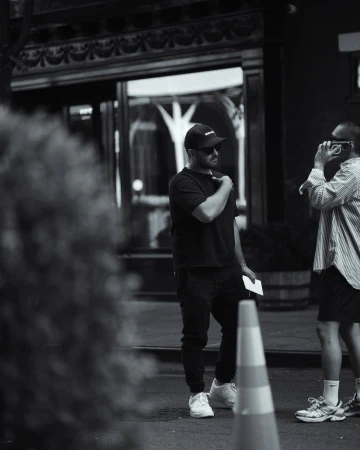 Man wearing a dark t-shirt and baseball cap standing outdoors next to another person holding a phone, shot by Rayan Ayash.
