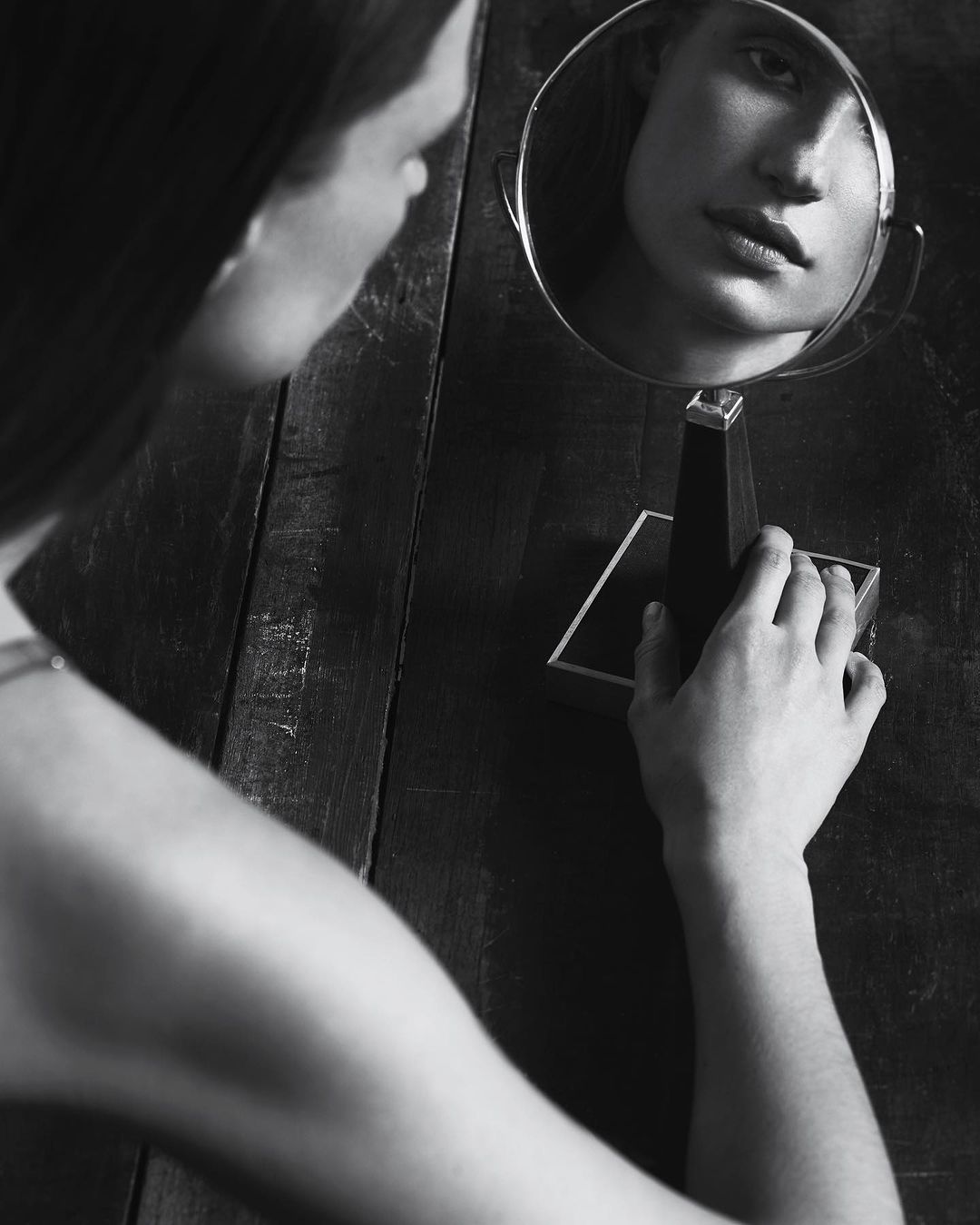 Black and white portrait of a woman looking into a small tabletop vanity mirror set on a dark wooden surface.