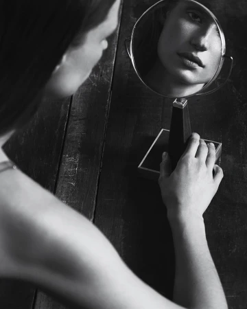 Black and white portrait of a woman looking into a small tabletop vanity mirror set on a dark wooden surface.