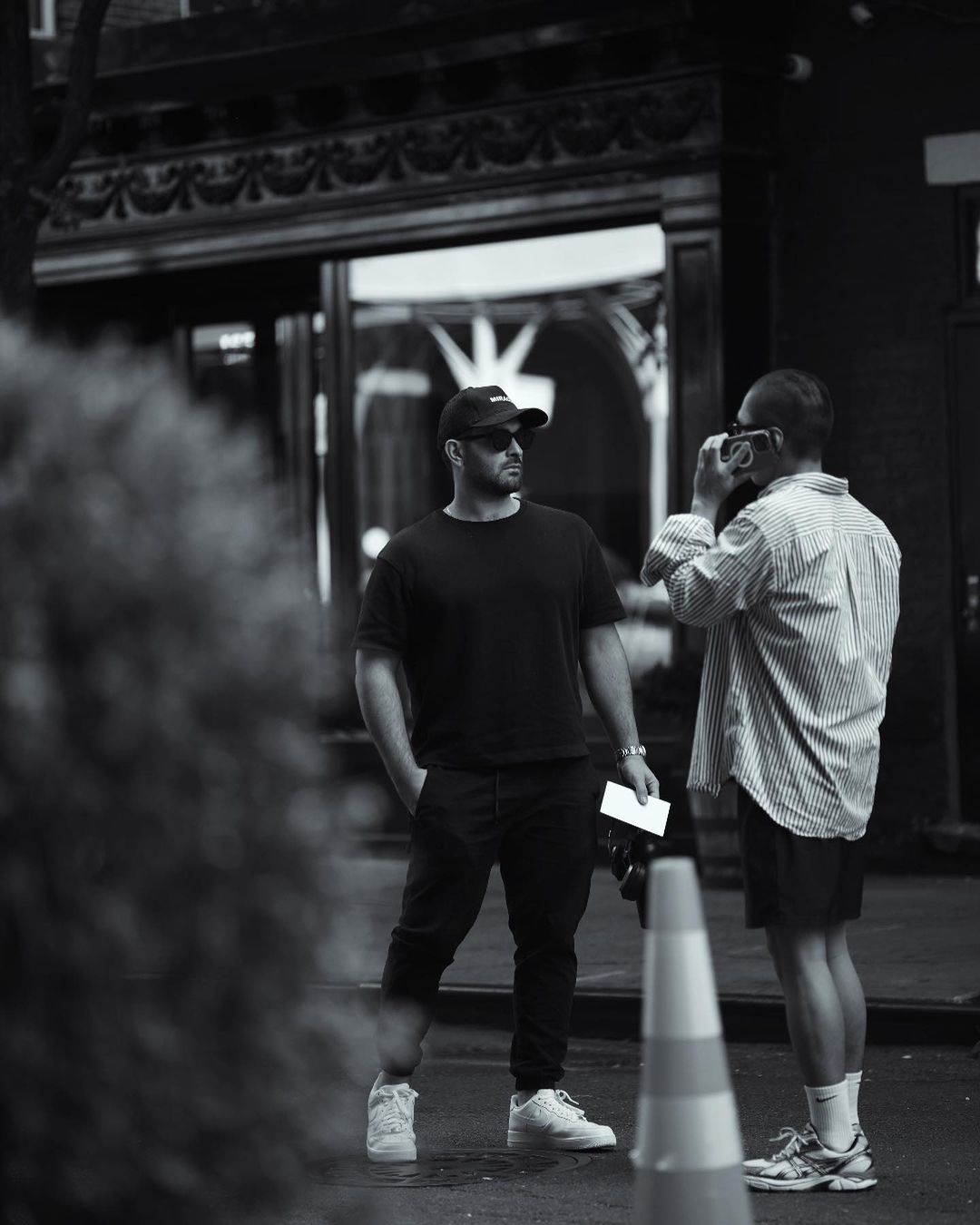 Two men stand outside a building. One man is holding a camera. The other man is wearing a baseball cap and sunglasses.