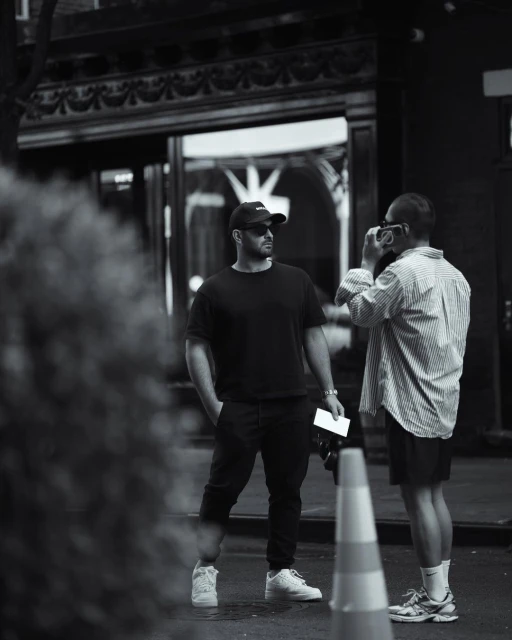 Two men stand outside a building. One man is holding a camera. The other man is wearing a baseball cap and sunglasses.