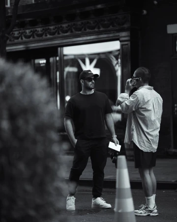 Two men stand outside a building. One man is holding a camera. The other man is wearing a baseball cap and sunglasses.