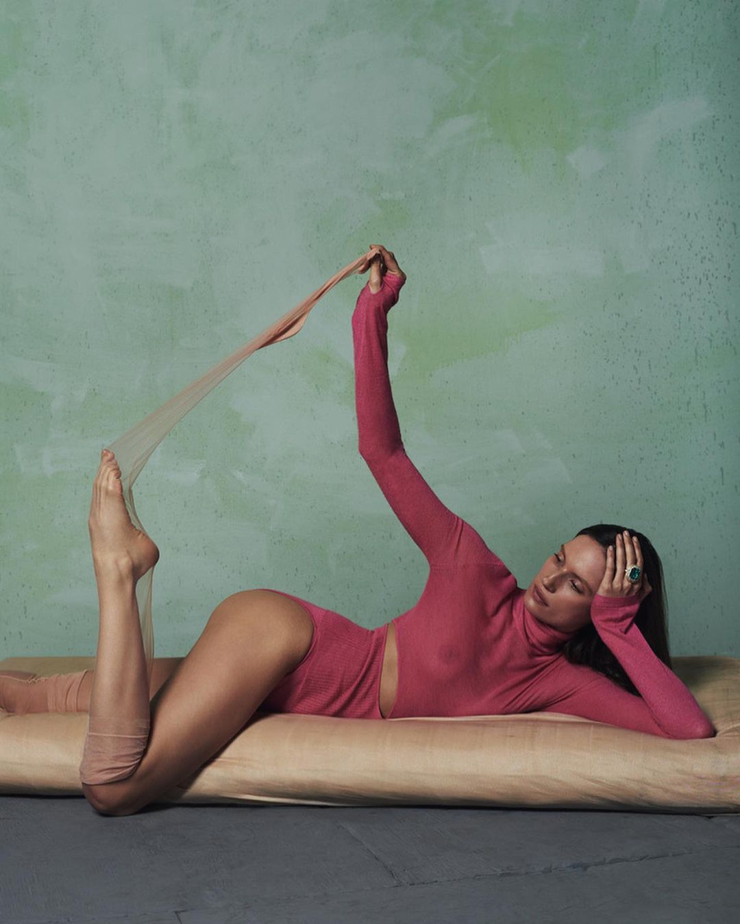 Adult female in a pink bodysuit stretches out on a beige cushion against a textured green background in a fashion or beauty shot.