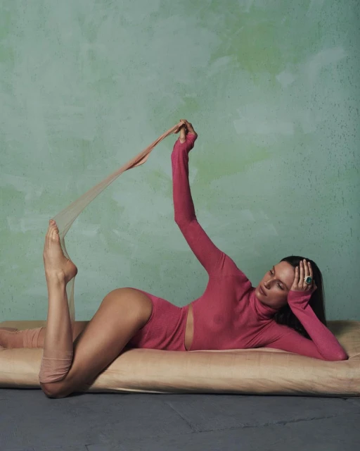 Adult female in a pink bodysuit stretches out on a beige cushion against a textured green background in a fashion or beauty shot.
