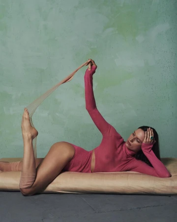 Adult female in a pink bodysuit stretches out on a beige cushion against a textured green background in a fashion or beauty shot.