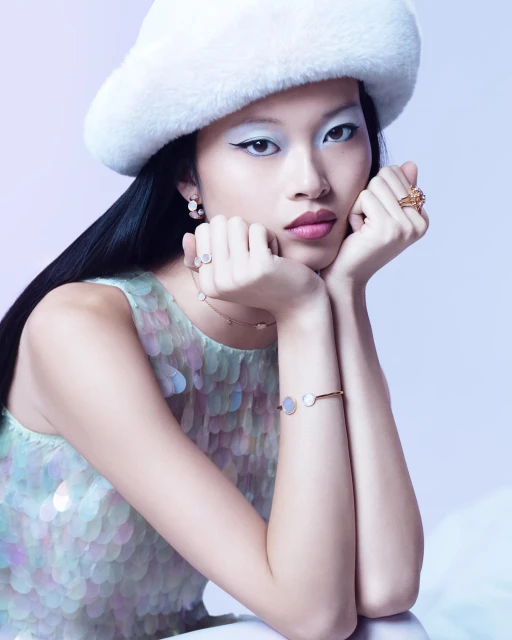 Asian woman with white eyeshadow and a fluffy white hat poses with jewelry, wearing a sparkling dress in a light-filled indoor studio.