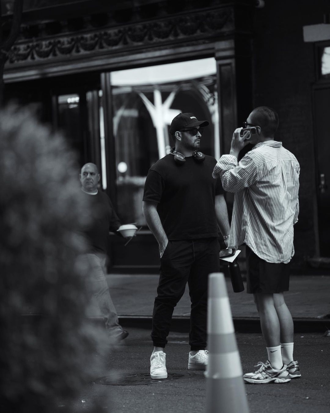 Black and white shot of two men in an urban street setting; one holds a camera as a third man walks by.