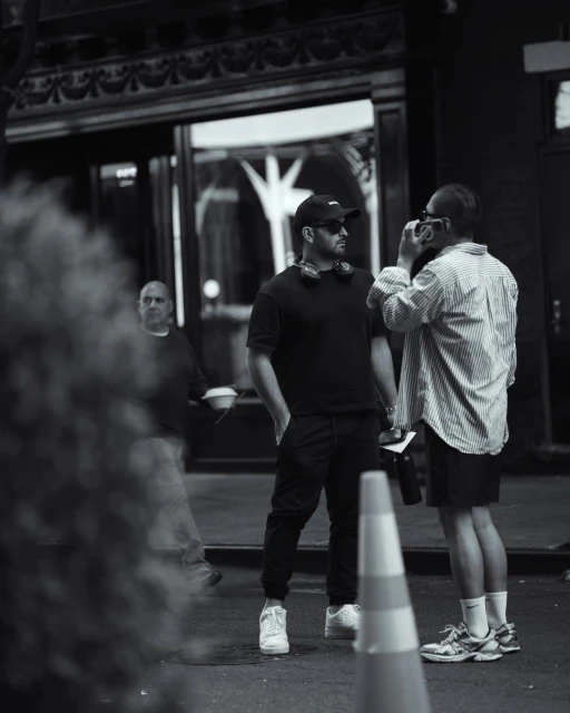 Black and white shot of two men in an urban street setting; one holds a camera as a third man walks by.