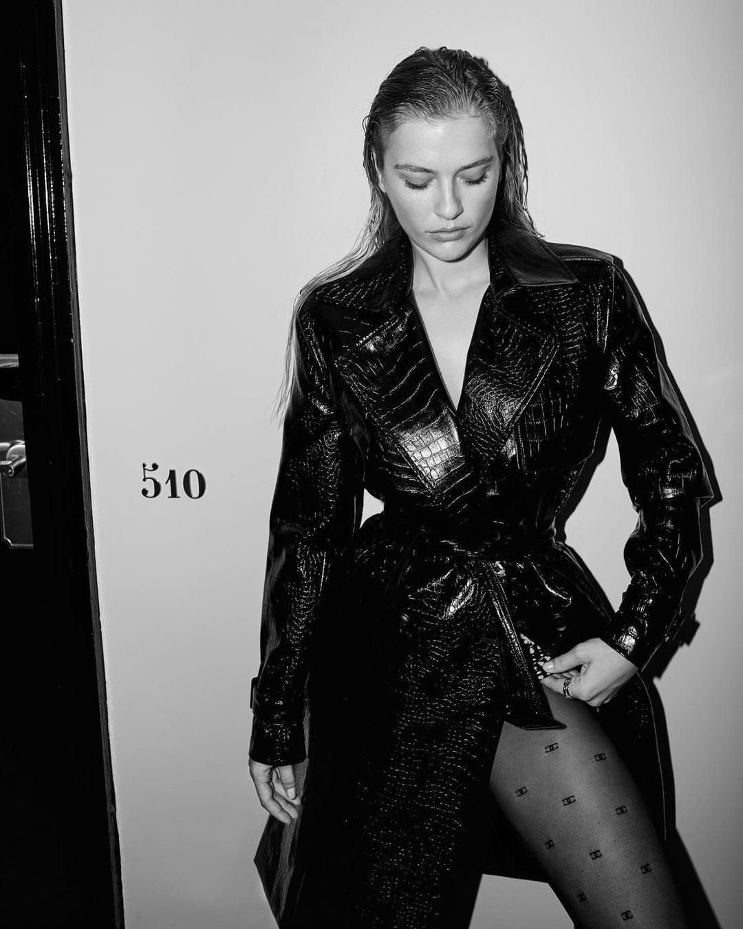 Black and white portrait of a woman in a crocodile-print coat and patterned tights looking down, posed against a light wall with a door.