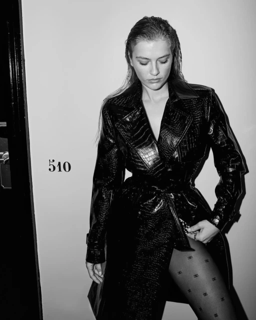 Black and white portrait of a woman in a crocodile-print coat and patterned tights looking down, posed against a light wall with a door.
