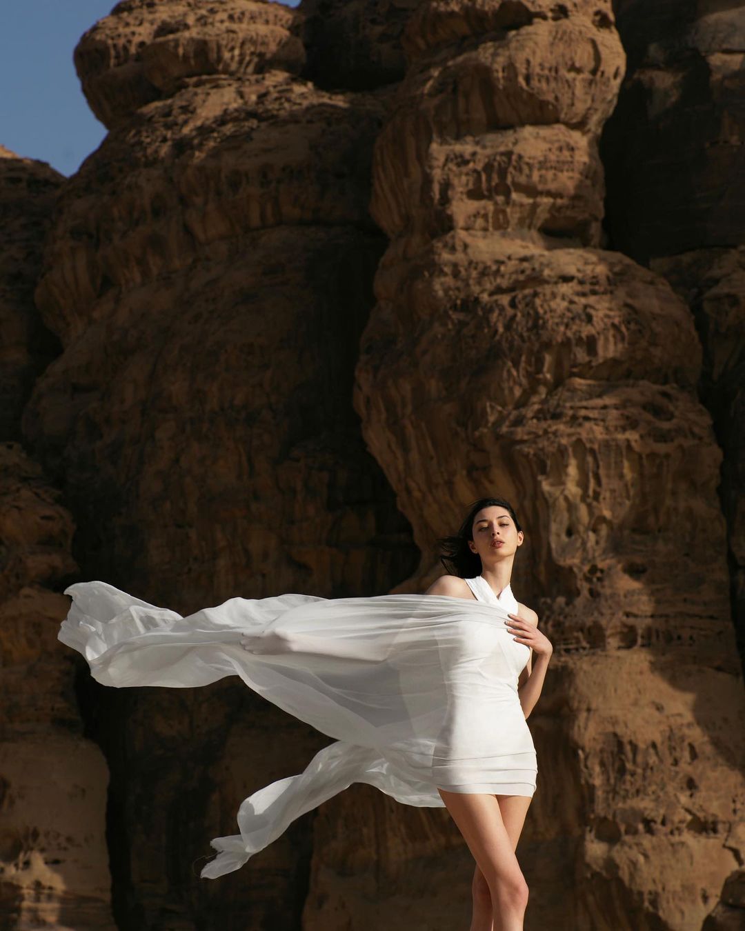 Woman in a white dress posing in front of a large rock formation on a sunny day, a sheer cloth billowing around her.