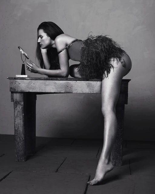 Monochrome portrait of a woman leaning on a rustic wooden table, gazing into a small mirror, wearing a feathered garment. Studio setting.