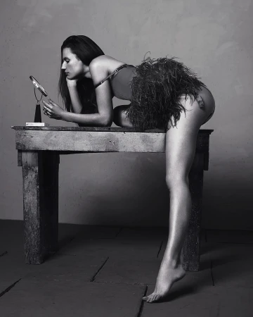Monochrome portrait of a woman leaning on a rustic wooden table, gazing into a small mirror, wearing a feathered garment. Studio setting.