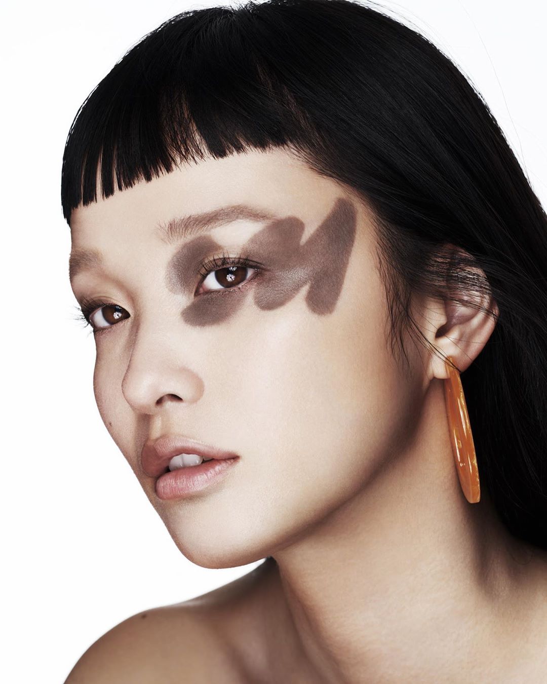 Close-up portrait of a stylish Asian woman with bangs, wearing oversized orange earrings, and bold makeup, set against a clean, white background.