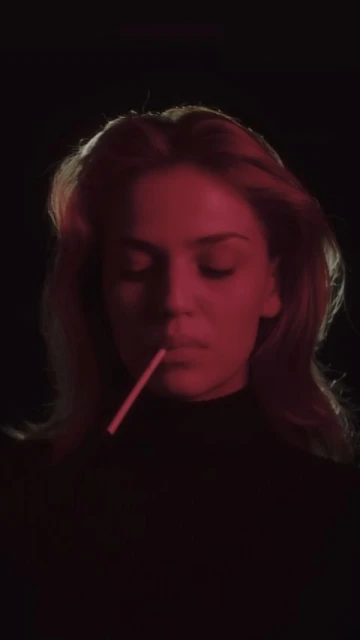 Portrait of a woman with light hair holding a lollipop stick in her mouth against a dark background with strong red lighting.