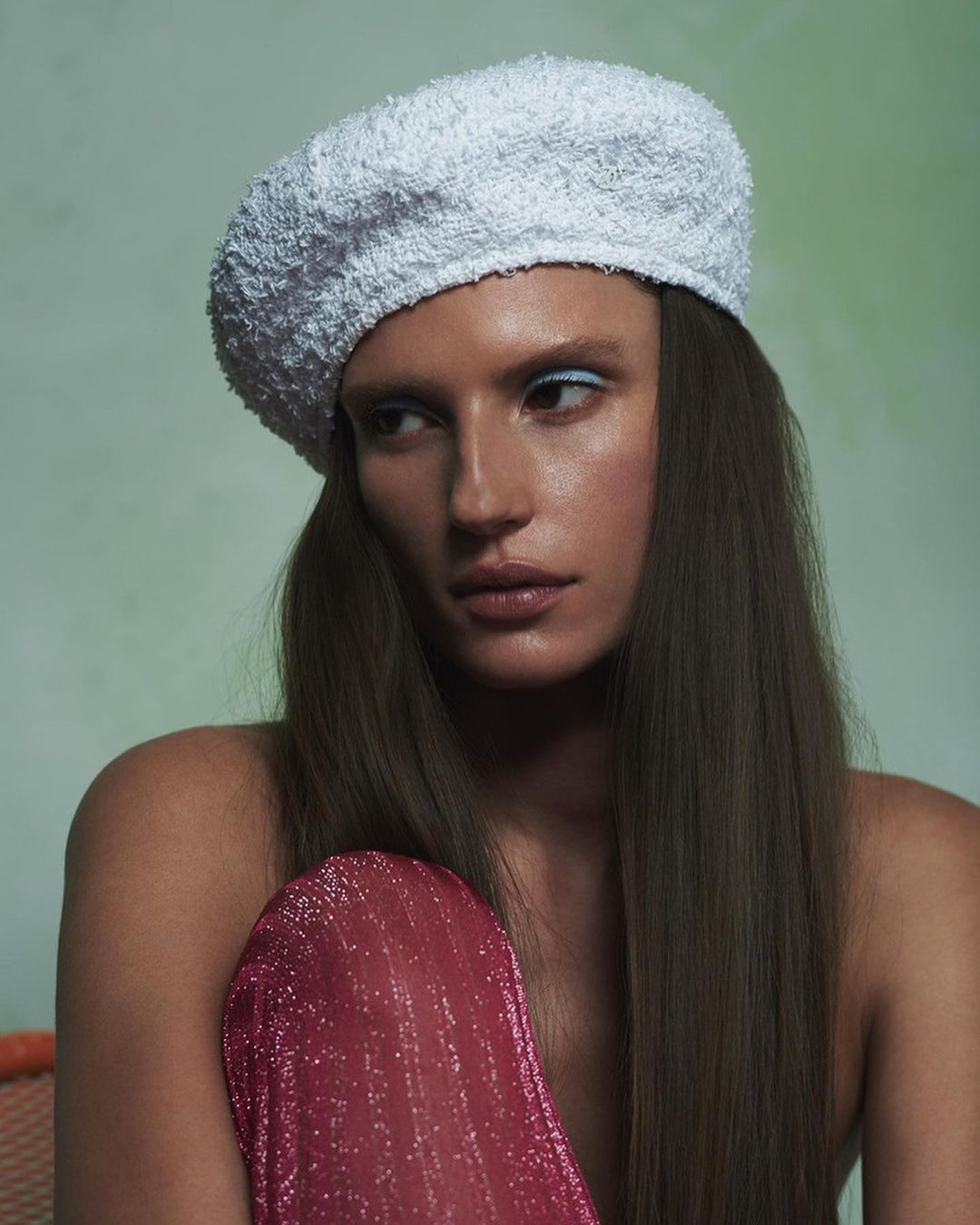 Portrait of a fair-skinned young woman wearing a beret and shoulder-revealing shiny pink top, posed against a soft green background.