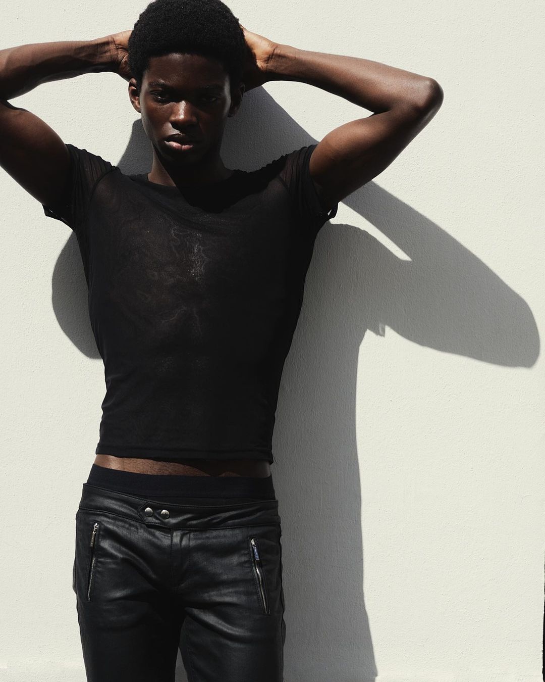 Portrait of Black man in black mesh shirt and leather pants posing against white wall with arm raised above his head casting shadow.