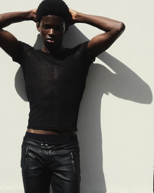 Portrait of Black man in black mesh shirt and leather pants posing against white wall with arm raised above his head casting shadow.