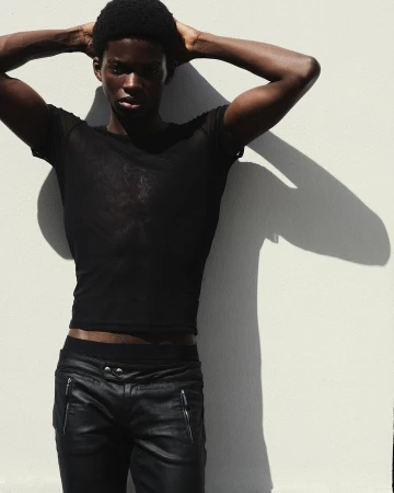 Portrait of Black man in black mesh shirt and leather pants posing against white wall with arm raised above his head casting shadow.