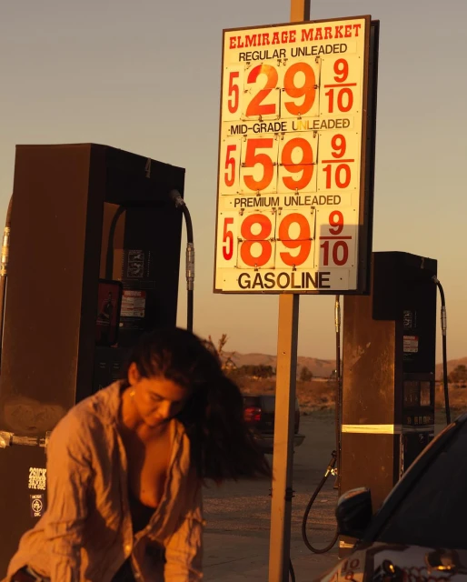 Woman standing near a vintage gas pump at sunset with a price sign displaying $5.29 for regular unleaded gas, in an outdoor setting.