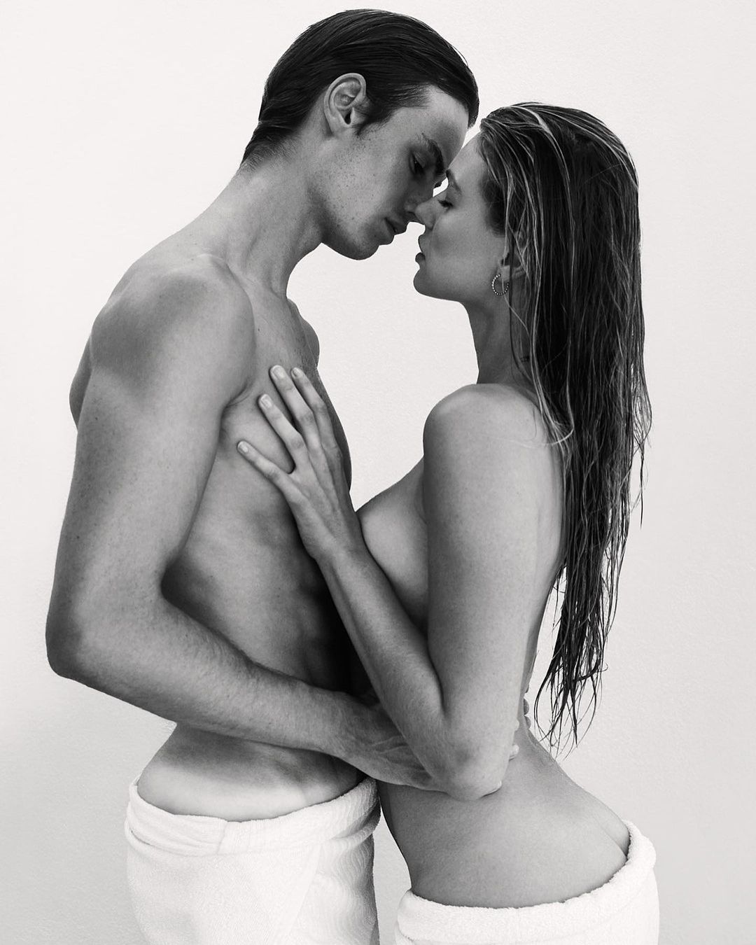 Striking black and white portrait of a couple embracing intimately, both wearing white towels, set against a clean, light backdrop.