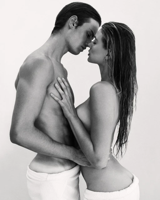 Striking black and white portrait of a couple embracing intimately, both wearing white towels, set against a clean, light backdrop.