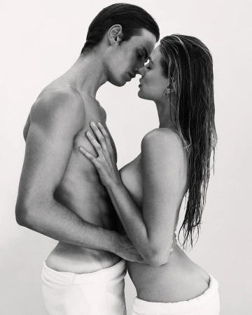 Striking black and white portrait of a couple embracing intimately, both wearing white towels, set against a clean, light backdrop.