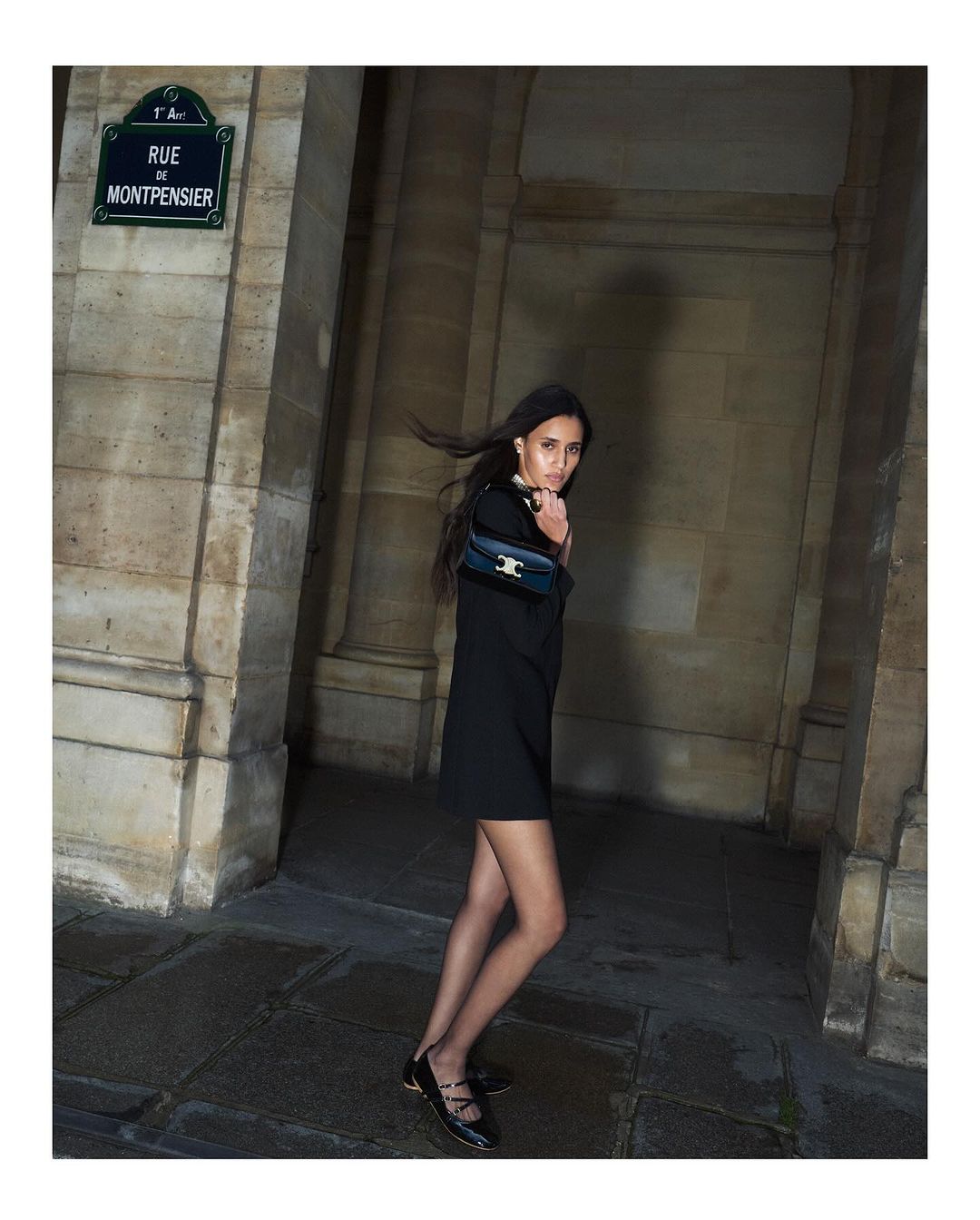 Woman in a black dress carrying a shoulder bag walking near stone architectural arches at night.