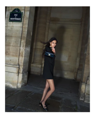 Woman in a black dress carrying a shoulder bag walking near stone architectural arches at night.