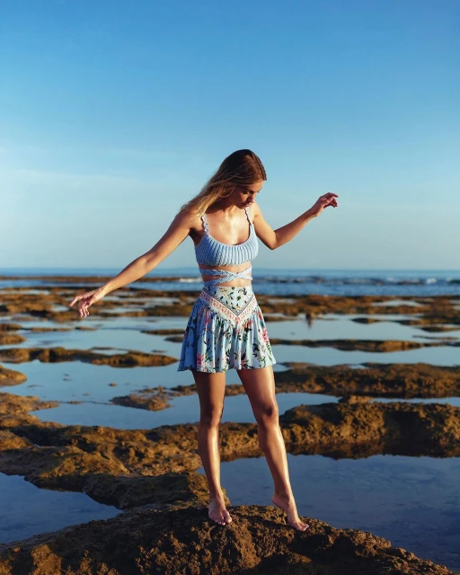 Blonde woman in floral skirt and matching crop top stands barefoot on rocks with arms outstretched on a sunny beach.