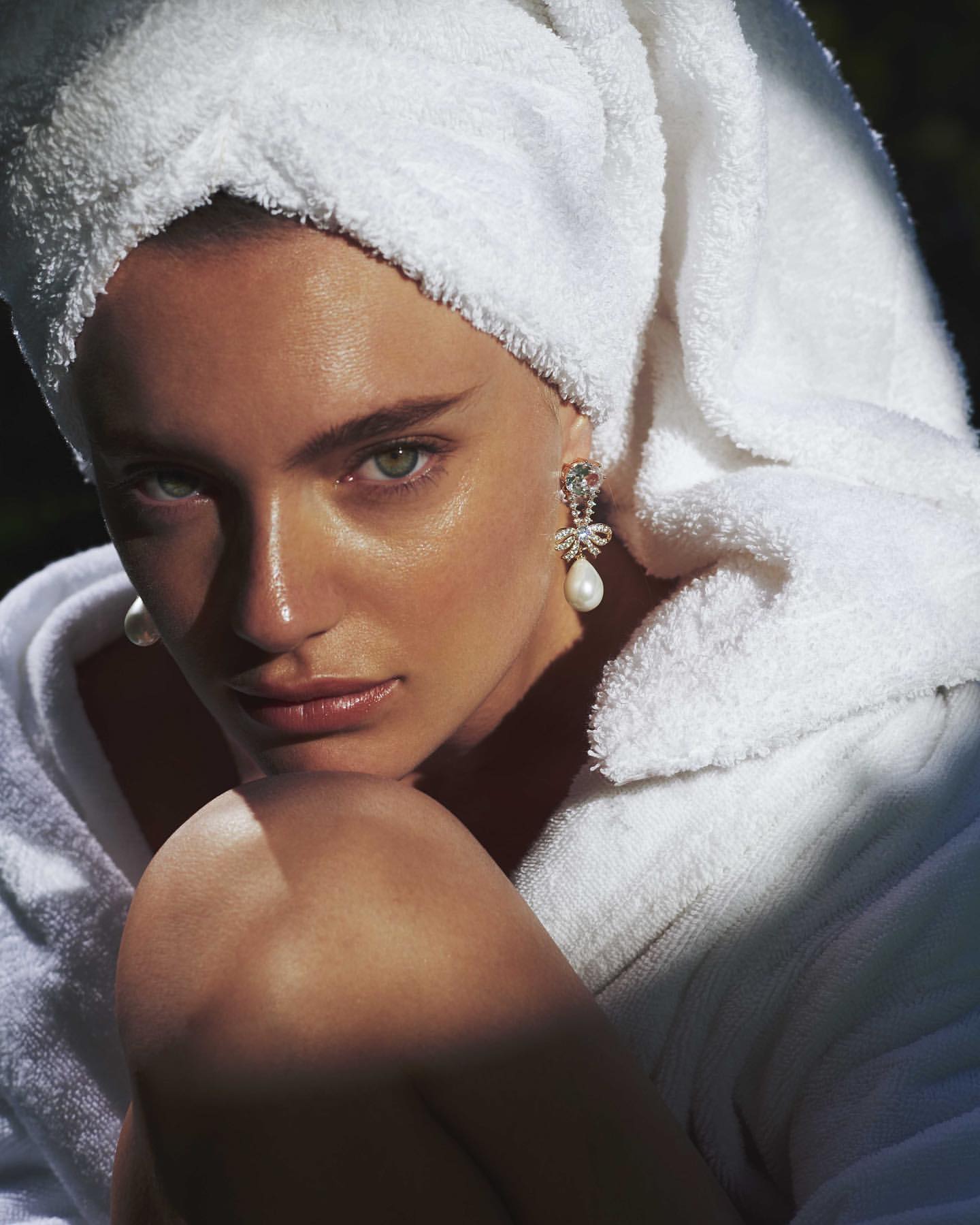 Close-up portrait of a woman with fair skin, wearing a white towel on her head and large pearl earrings, looking at the camera.