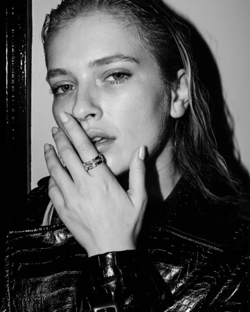 Close-up portrait of a woman with wet-look hair wearing a shiny leather jacket, holding her hand up to her face with a ring visible on her finger.