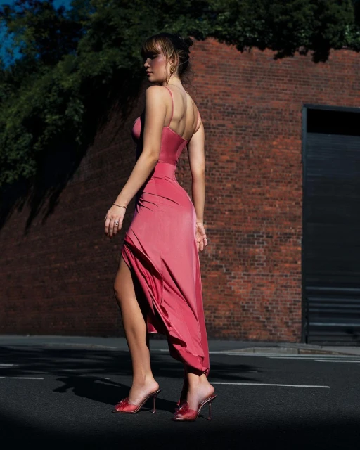 A woman in a pink dress and high heels walks on an asphalt road next to a red brick wall, with greenery above and shadows on the ground.