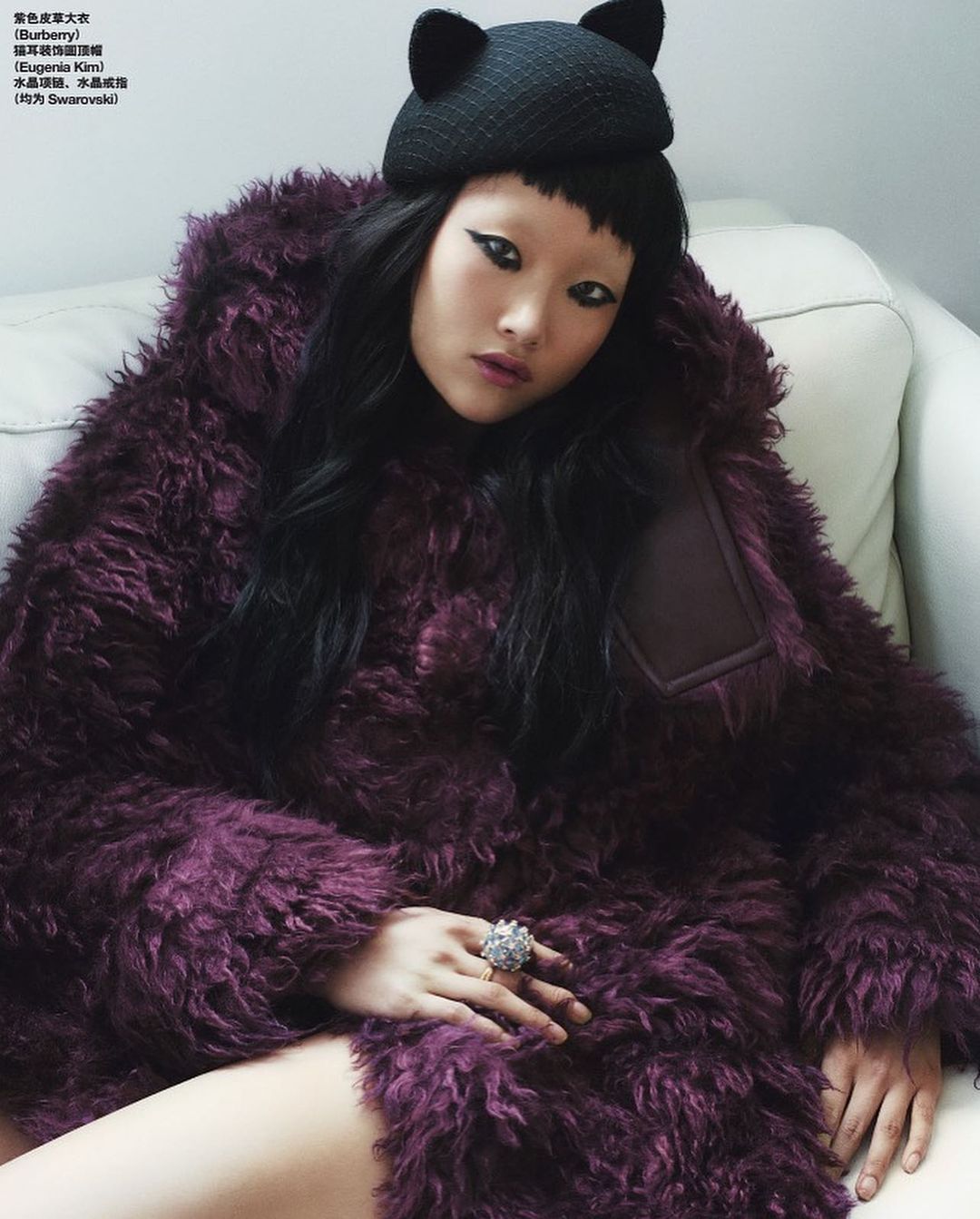 Woman wearing a furry purple coat and a black knit cat-ear hat poses indoors with dramatic makeup, long black hair, and an oversized ring.