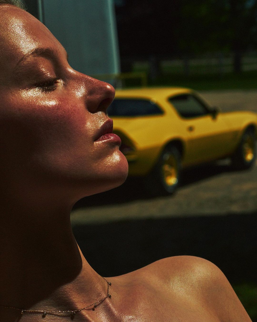 Close-up of a model in profile with glossy skin, sunlight, and a gold necklace. A blurred yellow car is in the background.