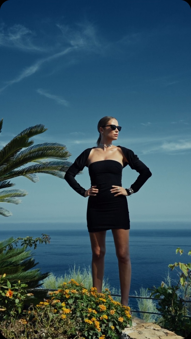 A woman in a black dress stands with hands on hips against a backdrop of the ocean and sky with tropical vegetation in the foreground.