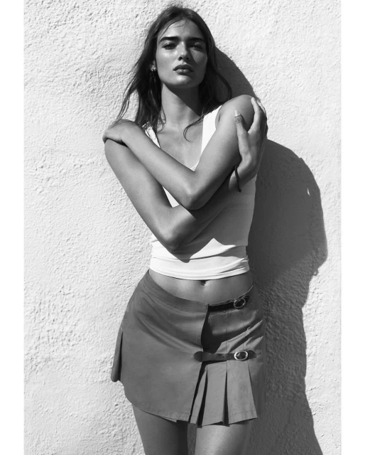 Monochrome portrait of a woman in a white top and short skirt posing in front of a textured wall in natural light.