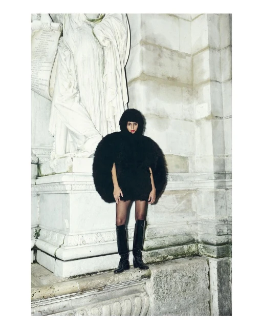 Fashion model poses in a round, black fur coat and knee-high boots against a neoclassical monument on an overcast day.