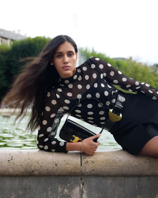 Adult woman in a polka dot top and black skirt poses outdoors with a black purse against a green background of trees and water.
