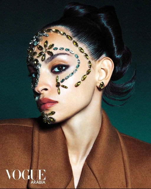 Close-up of a woman with dark hair wearing a brown coat and face gems in a fashion advertisement for Vogue Arabia magazine.
