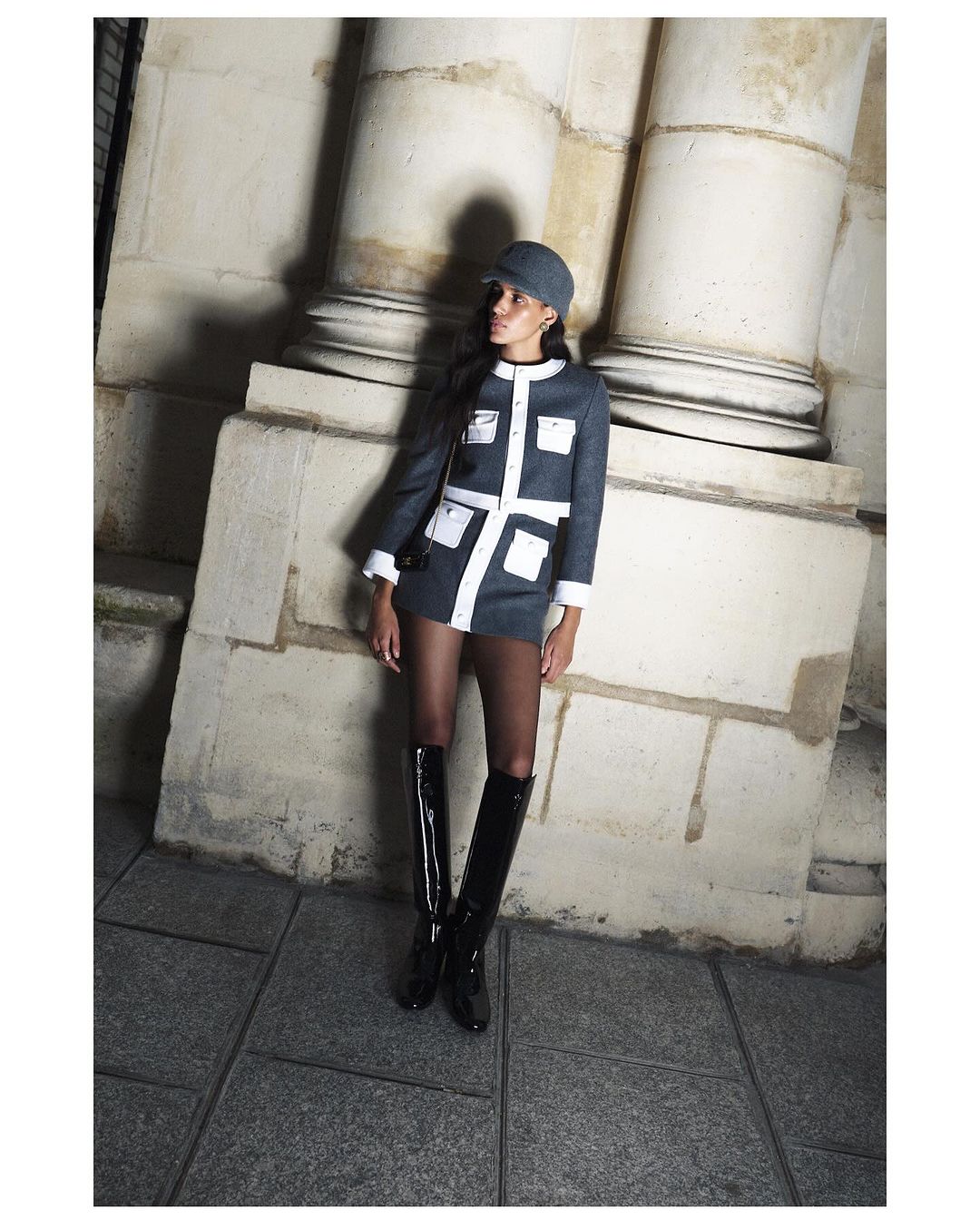 Elegant woman in a short blue and white dress with matching hat posing near stone columns in shiny black boots. Fashion photography.