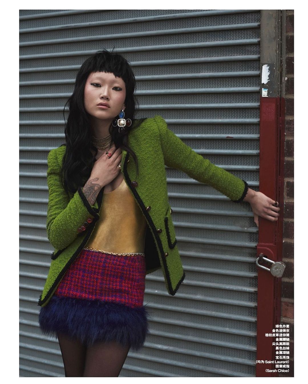 A stylish Asian woman poses in front of a roll-up door, wearing a textured green jacket, gold top, and multicolored miniskirt with dark hair.