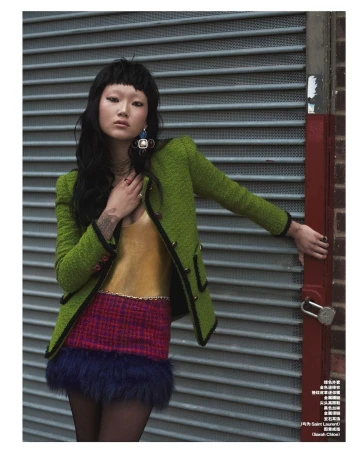 A stylish Asian woman poses in front of a roll-up door, wearing a textured green jacket, gold top, and multicolored miniskirt with dark hair.