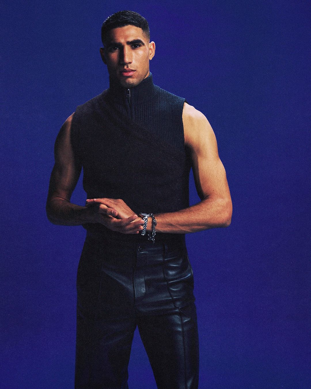 Portrait of a stylish adult male wearing a sleeveless black top and shiny black pants standing in front of a deep blue background.