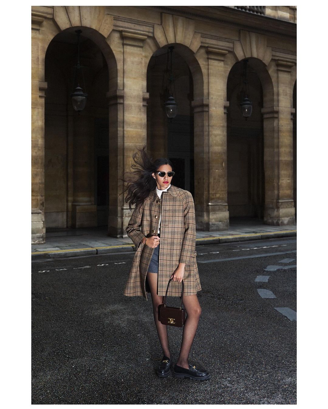 A fashionable woman stands on a city street wearing a plaid coat, black tights, and loafers while holding a small purse in a wide shot.