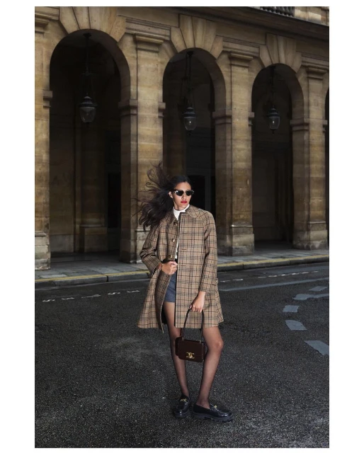A fashionable woman stands on a city street wearing a plaid coat, black tights, and loafers while holding a small purse in a wide shot.