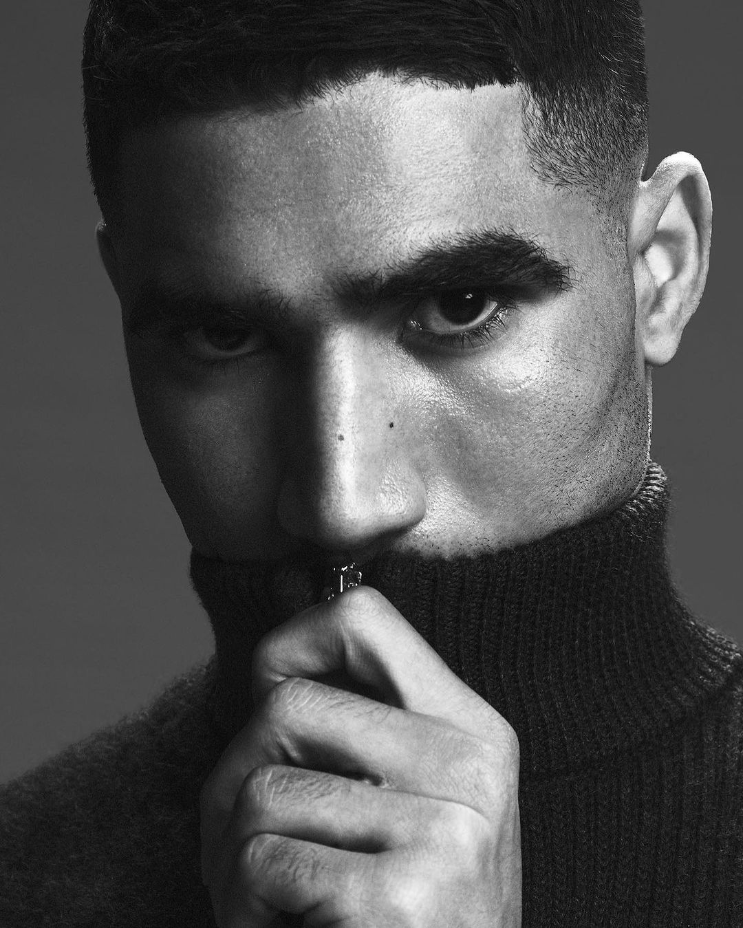 Close-up black and white portrait of a young adult male with a buzz cut, wearing a turtleneck sweater zipped up to his mouth.