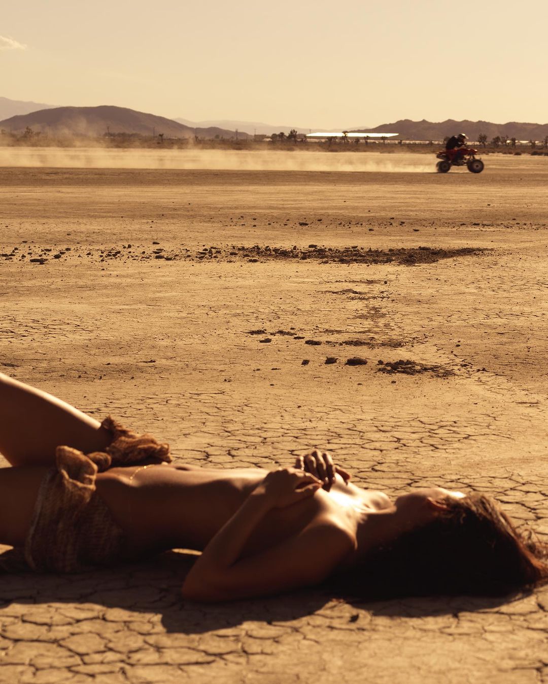 Woman lies on cracked earth as an ATV drives in the distance, with a desert landscape, and clear sky in a warm color grading.