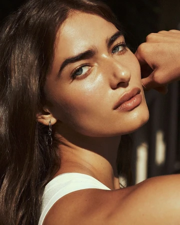 Close-up portrait of a woman with glowing skin wearing a white top and small hoop earrings, photographed by Rayan Ayash.