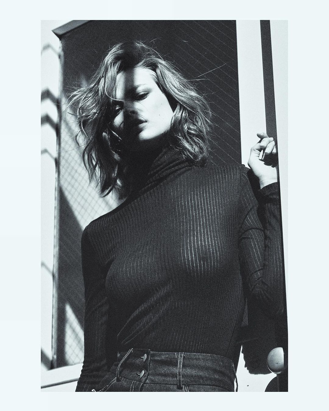 Black and white portrait of a woman with shoulder-length wavy hair wearing a black turtleneck and jeans, posing near a window.